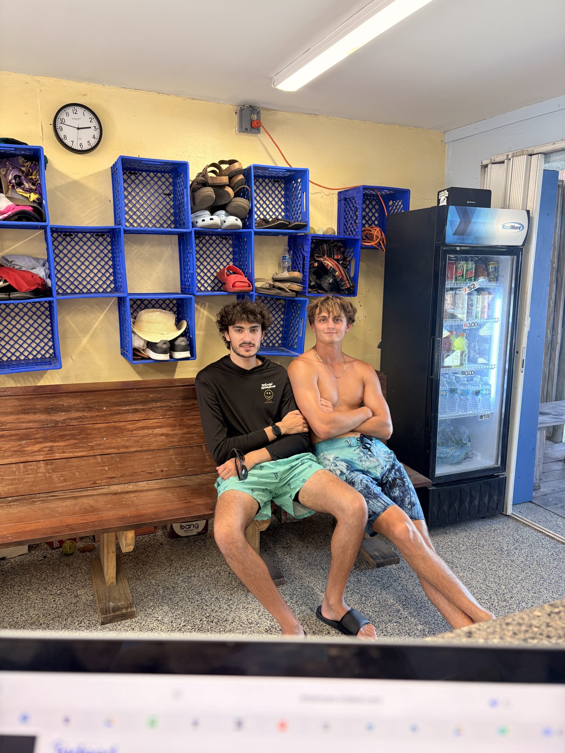 Two men sitting in a room with blue cubbies and a fridge in the background.