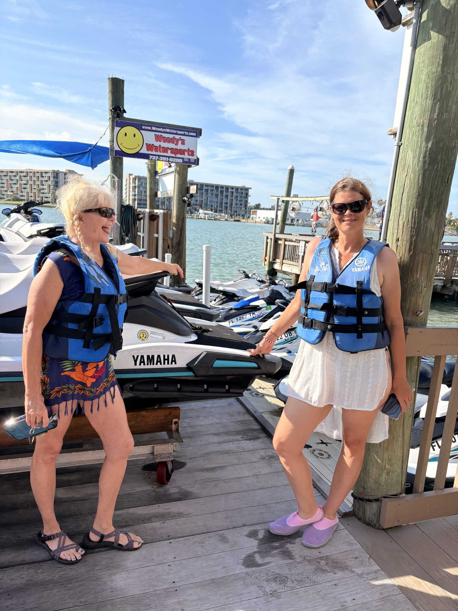 Two women in life jackets standing on a dock with jet skis.