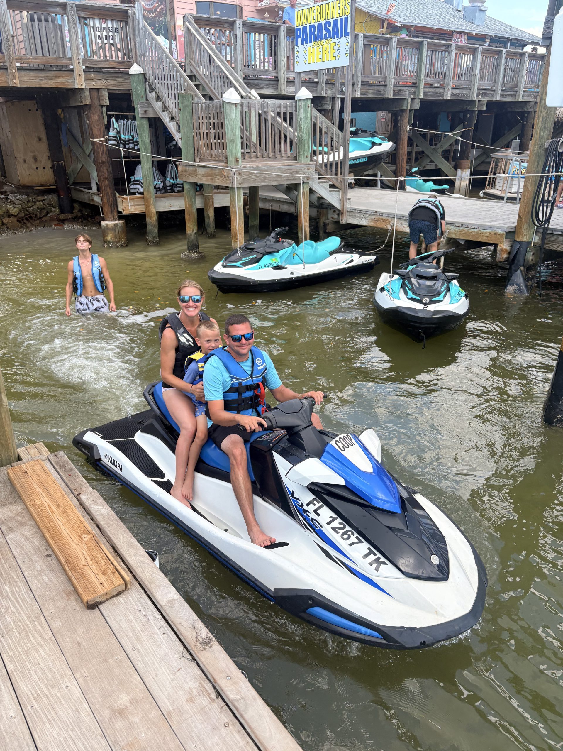 Two people on a jet ski by a dock, another person standing nearby in shallow water.