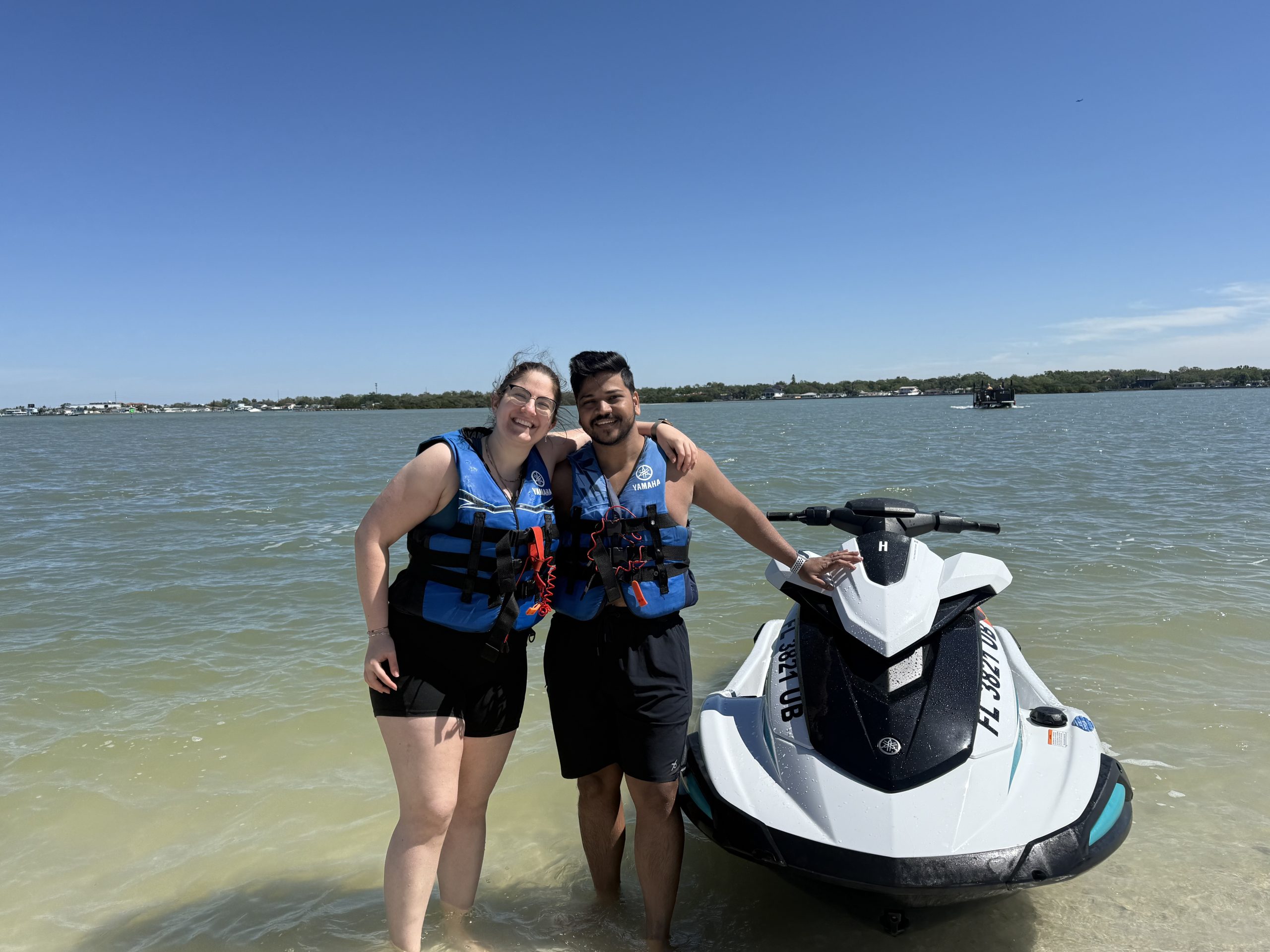 Two people in life jackets stand by a jet ski on a sunny beach.