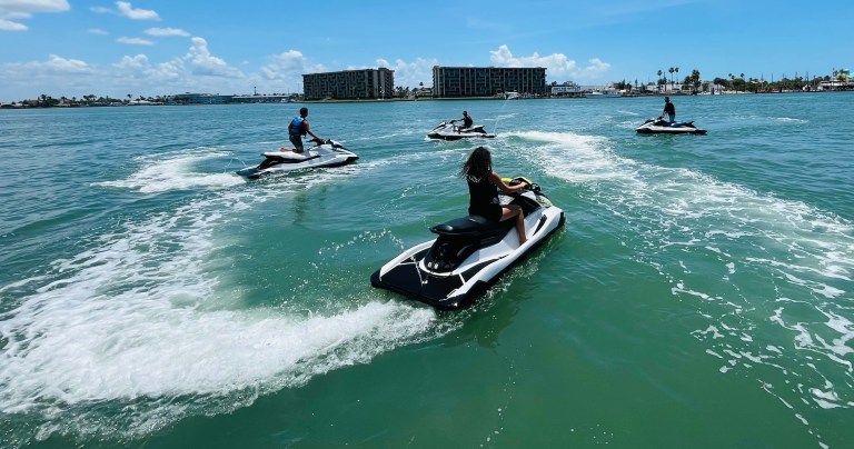 Four people riding jet skis on a sunny day with buildings in the background.