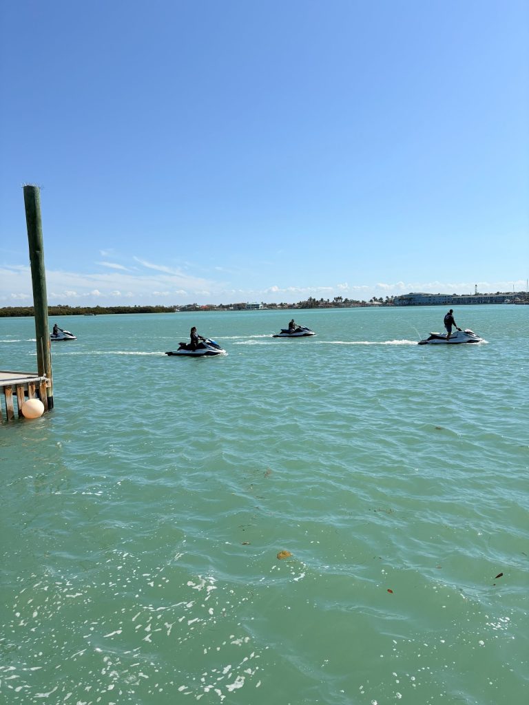 People riding jet skis on calm blue water, with a clear sky above.